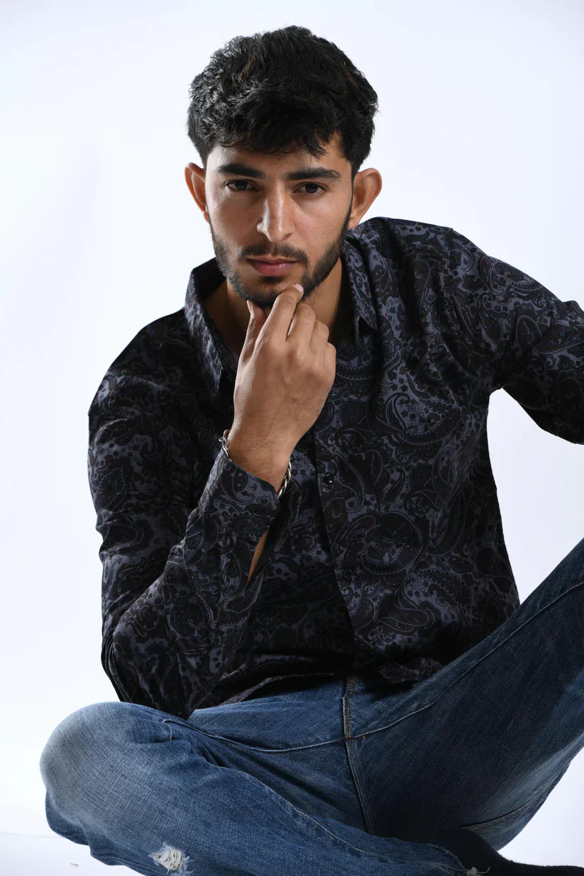 Man wearing a black printed shirt with grey patterns, sitting casually.