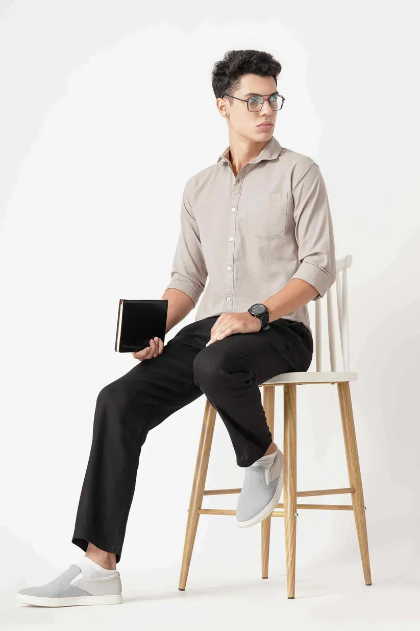 Man wearing a beige linen shirt with black pants, holding a notebook, sitting on a stool.