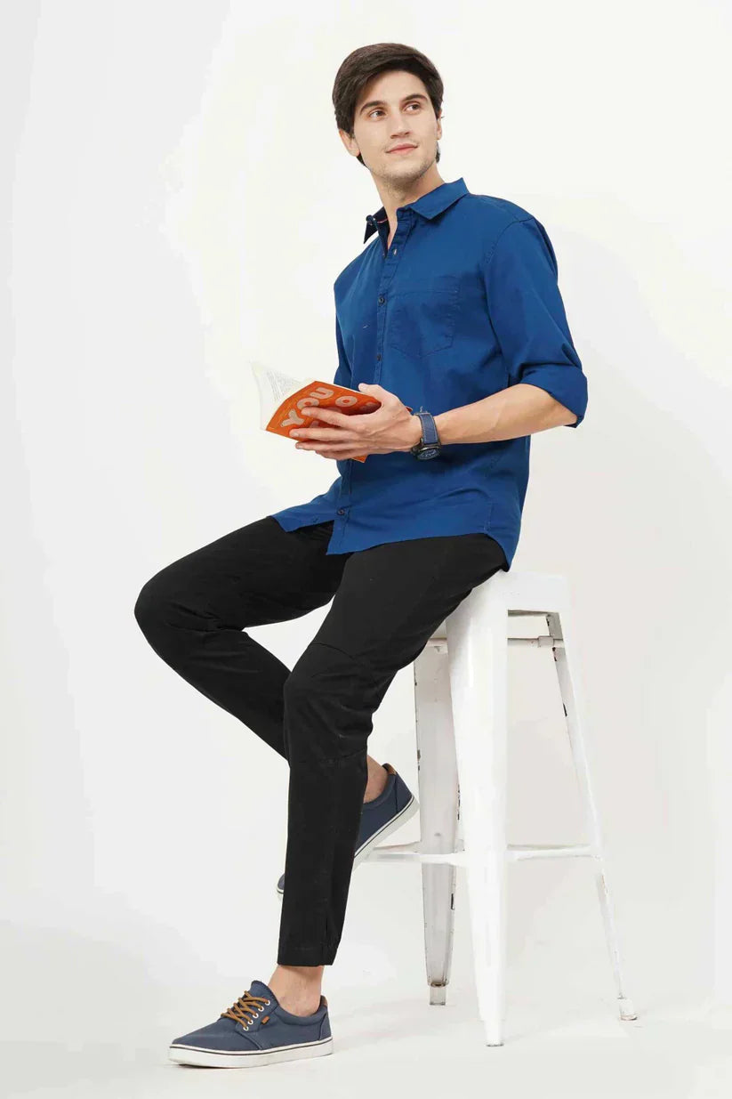 Man wearing a stylish blue plain shirt, sitting on a stool, holding a book.