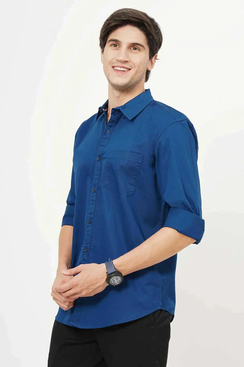 A smiling man in a blue plain shirt adjusting his sleeve.