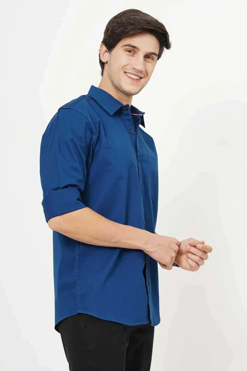 A smiling man in a blue plain shirt adjusting his sleeve.